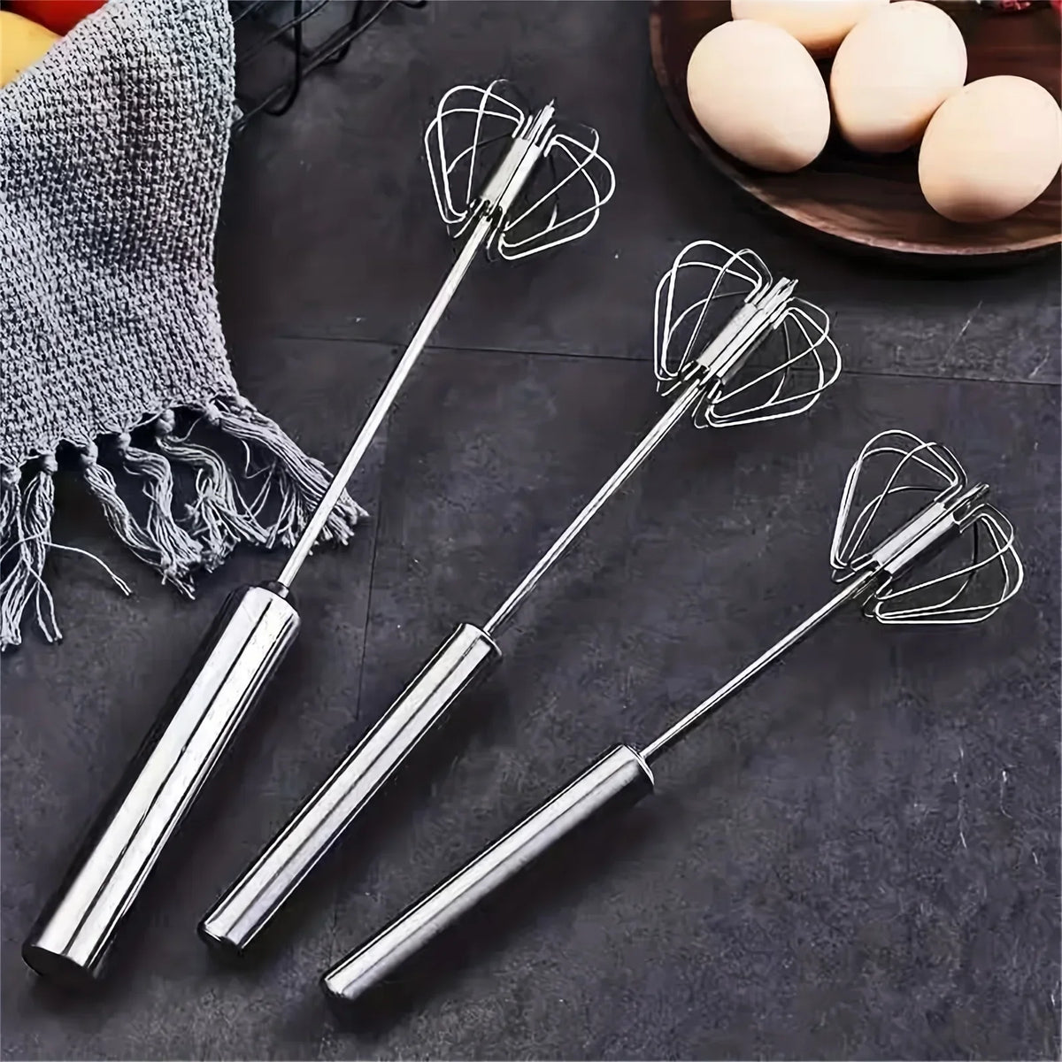 Three stainless steel handheld kitchen whisks on dark countertop with a gray towel and wooden bowl of eggs