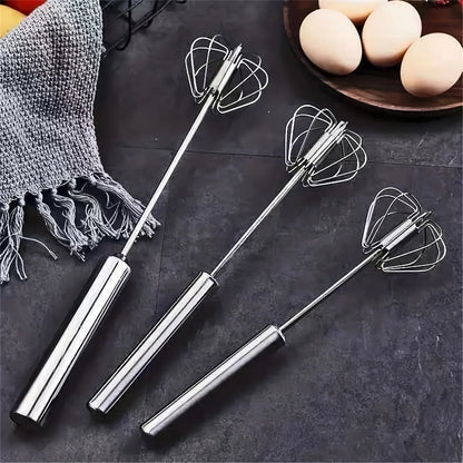 Three stainless steel handheld kitchen whisks on dark countertop with a gray towel and wooden bowl of eggs