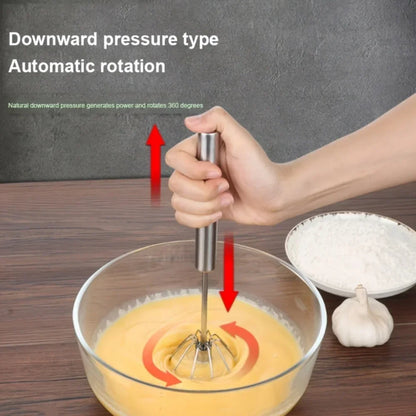 Hand using a stainless steel rotary whisk to mix batter in a glass bowl on wooden table with flour and garlic nearby