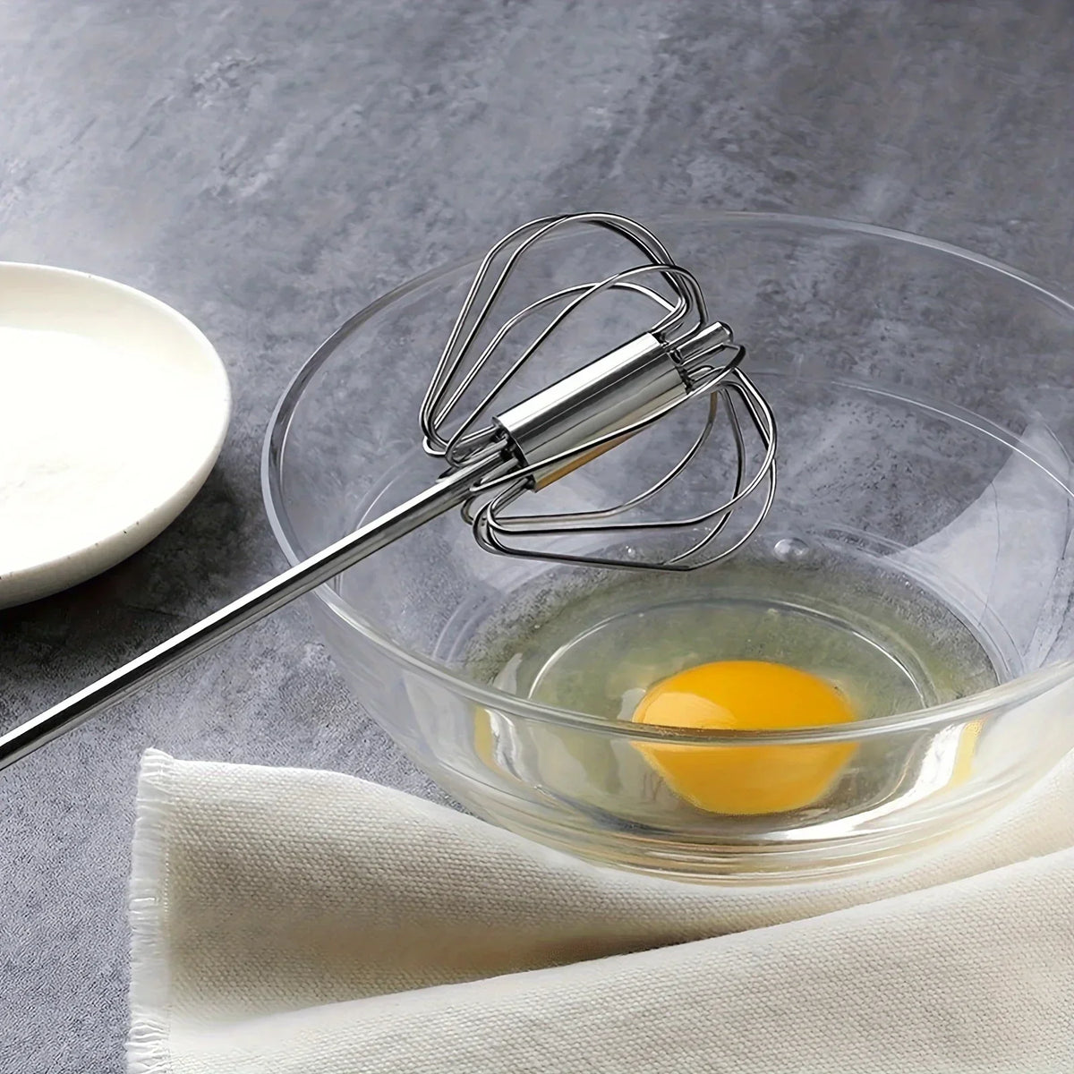 Glass bowl with cracked egg yolk and stainless steel hand whisk on grey surface with white cloth
