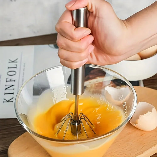 Hand using stainless steel whisk to beat eggs in glass bowl on wooden board with broken eggshells