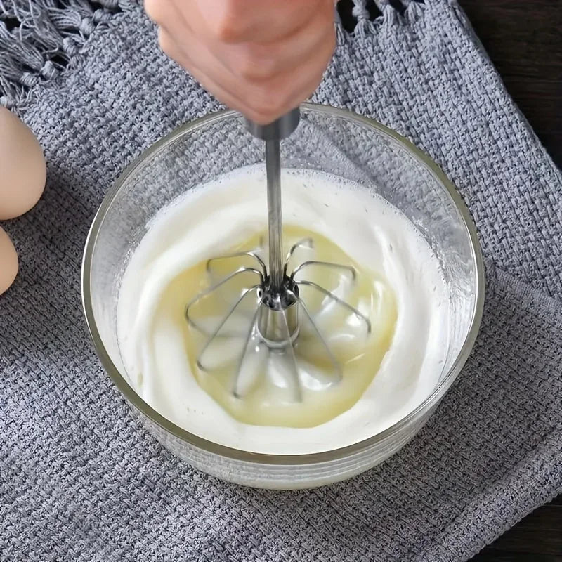 Hand using whisk to beat egg whites in glass bowl on gray knitted cloth with eggs nearby