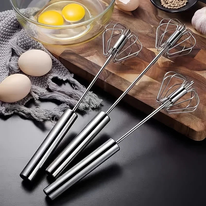Three stainless steel egg whisks on black surface near eggs, cracked eggs in bowl, cutting board, and spices