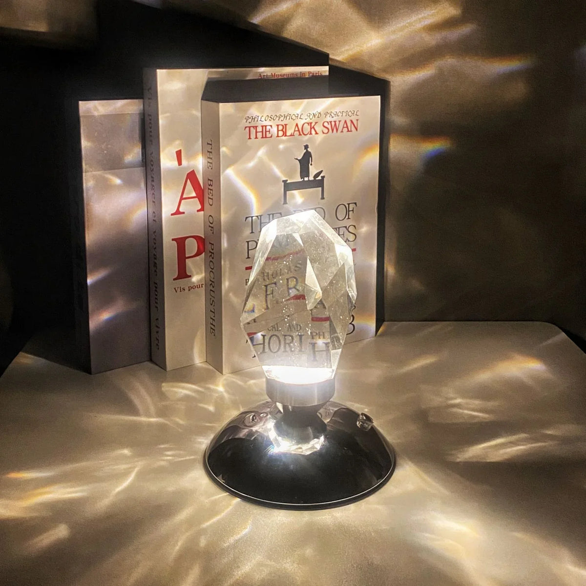 Crystal prism lamp casting light patterns on surface near books including The Black Swan